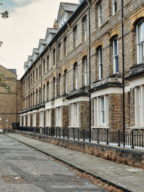 A row of three-storey Victorian residential buildings constructed from yellow-brown brick, featuring white-framed sash windows and small front yards enclosed by black metal railings. The pavement along the buildings is made of stone slabs, with a narrow, slightly worn asphalt street in front. The scene is illuminated by natural daylight, with some fallen leaves visible on the ground. The architecture and surroundings suggest an urban setting in a historic area, with the image associated with the service page for Abbeville Road end of tenancy cleaning checklist by Carpet Cleaning Clapham, highlighting surface cleaning and general hygiene in residential properties.