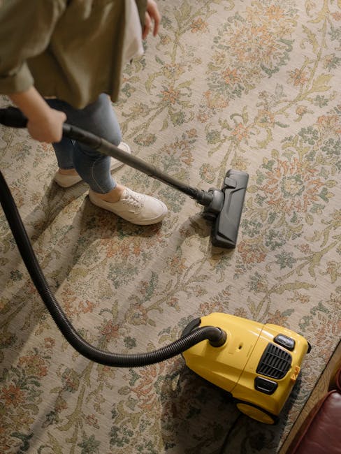 A person cleaning a traditional patterned area rug on a wooden floor using a yellow industrial vacuum cleaner with a black hose, demonstrating surface cleaning and maintenance in a living room setting. The person is dressed in casual clothing and light-colored sneakers, operating the vacuum over the beige and multicoloured floral carpet, with natural ambient lighting highlighting the clean and well-maintained condition of the rug. The scene emphasizes domestic cleaning practices relevant to avoiding common carpet cleaning mistakes, consistent with services provided by Carpet Cleaning Clapham.
