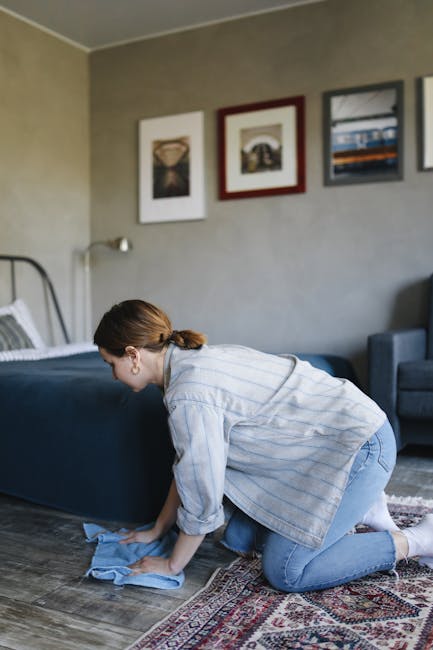 A woman wearing a light blue, striped shirt and jeans is kneeling on a patterned rug in a modern living room, carefully wiping a dark blue upholstered bed frame with a blue cloth. The room features a wooden floor, neutral-toned walls decorated with three framed pieces of artwork, and natural lighting coming from an off-screen window. A small wall-mounted lamp is positioned above the bed, and a grey armchair is visible in the background. The scene depicts domestic surface cleaning and maintenance, emphasizing the importance of thorough dusting and sanitisation, which is essential for avoiding common carpet cleaning mistakes in Clapham flats. Carpet Cleaning Clapham provides professional deep cleaning services to ensure homes are hygienic and well-maintained.