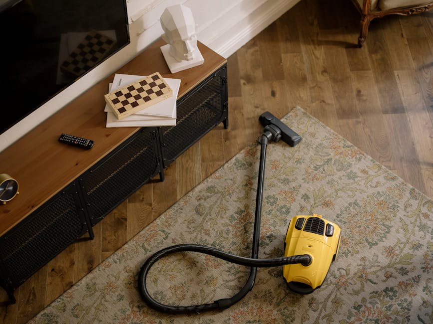 A person cleaning a traditional patterned area rug on a wooden floor using a yellow industrial vacuum cleaner with a black hose, demonstrating surface cleaning and maintenance in a living room setting. The person is dressed in casual clothing and light-colored sneakers, operating the vacuum over the beige and multicoloured floral carpet, with natural ambient lighting highlighting the clean and well-maintained condition of the rug. The scene emphasizes domestic cleaning practices relevant to avoiding common carpet cleaning mistakes, consistent with services provided by Carpet Cleaning Clapham.