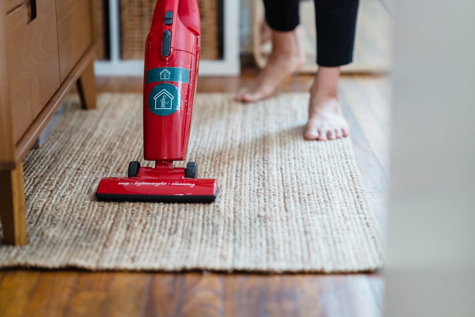 A person vacuuming a beige woven carpet in a residential room with wooden floors and a wooden cabinet on the left side. The individual is barefoot, and part of their black clothing is visible. The vacuum cleaner, which is red with blue control buttons, is positioned on the carpet, indicating surface cleaning. The room is well-lit, highlighting the clean and tidy condition of the carpet, and the scene suggests a routine household deep cleaning process. Carpet Cleaning Clapham offers professional domestic cleaning services including surface cleaning and sanitisation to maintain hygiene in living spaces.