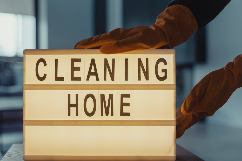 A person wearing orange cleaning gloves is adjusting a lightbox sign that displays the words 'CLEANING HOME' in bold black letters on a white background. The sign is placed on a table in a well-lit ro