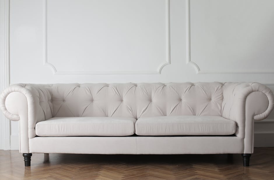A white tufted fabric sofa with rolled arms and dark wooden legs situated in a bright, minimalist room with white paneled walls. The sofa appears clean and well-maintained, with no visible stains or dust. The hardwood floor beneath the sofa is polished, adding to the room's tidy appearance. The lighting emphasizes the softness of the upholstery and highlights the neatness of the space, reflecting professional cleaning and surface sanitisation by Carpet Cleaning Clapham, specialising in domestic surface cleaning and deep cleaning services.