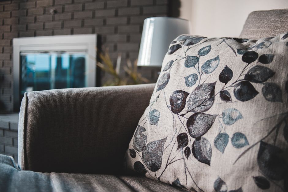 Close-up image of a beige fabric sofa with a decorative cushion featuring a leaf pattern in shades of blue and black, situated in a modern living room. The sofa's upholstery appears clean and well-maintained, with soft natural lighting highlighting its texture. In the background, there is a contemporary grey brick wall, a white electric fireplace with a glass front, and a sleek white table lamp emitting a gentle glow. The scene suggests a tidy, comfortable, and inviting domestic space, emphasizing surface cleaning and upholstery maintenance, aligning with services offered by Carpet Cleaning Clapham for deep cleaning and hygiene in busy homes.